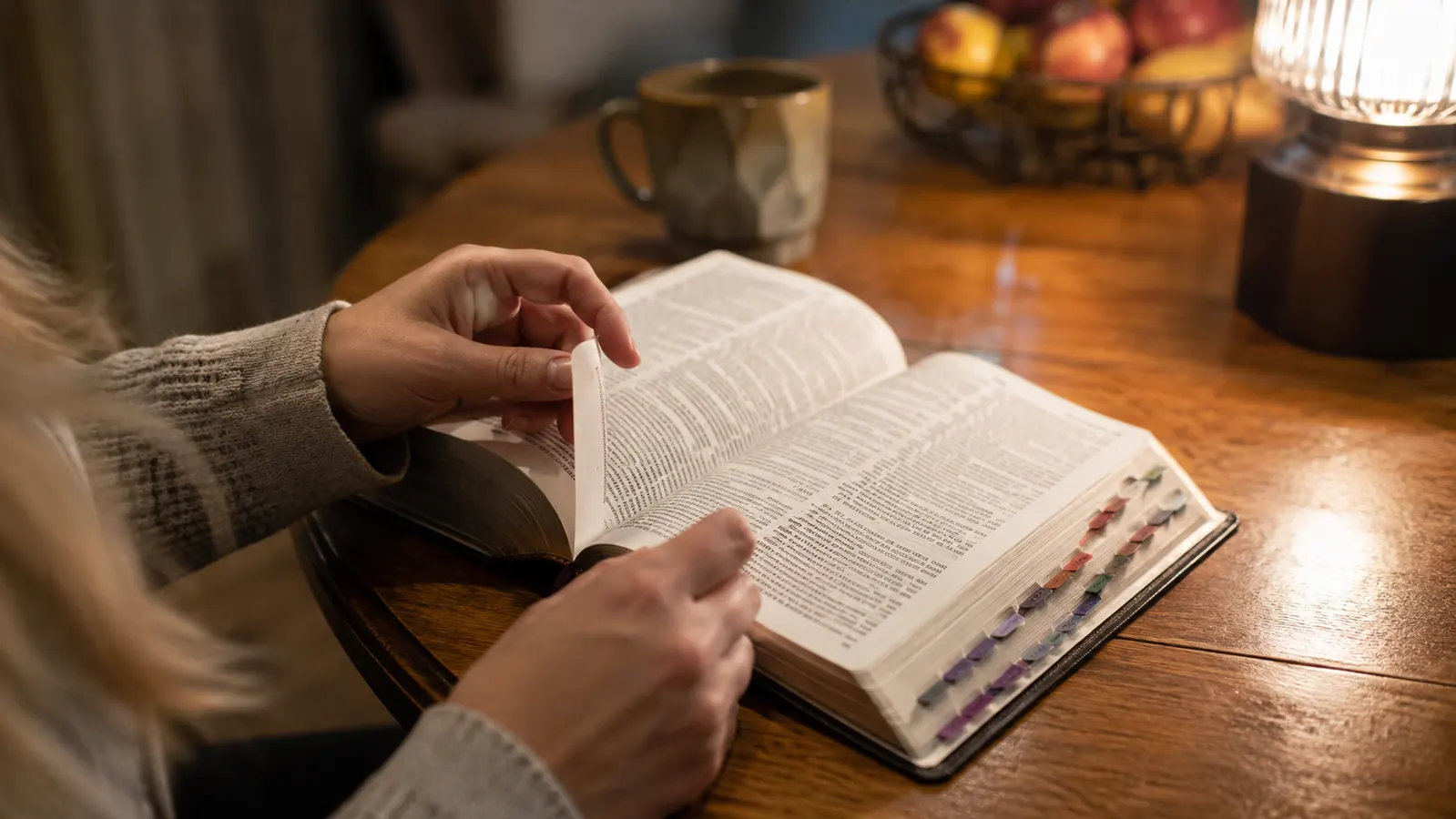 A woman's hands turning the pages of an open Bible with tabs along the page edge, warm lamp light on a kitchen table